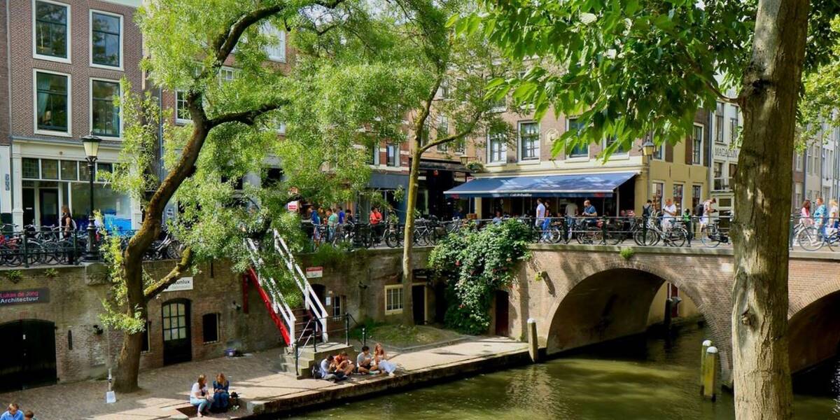 Link naar foto in Flickr: zicht op deel van de Oudegracht. Met mensen die aan de gracht zitten in de zon en mensen die bovenlangs de gracht lopen. 